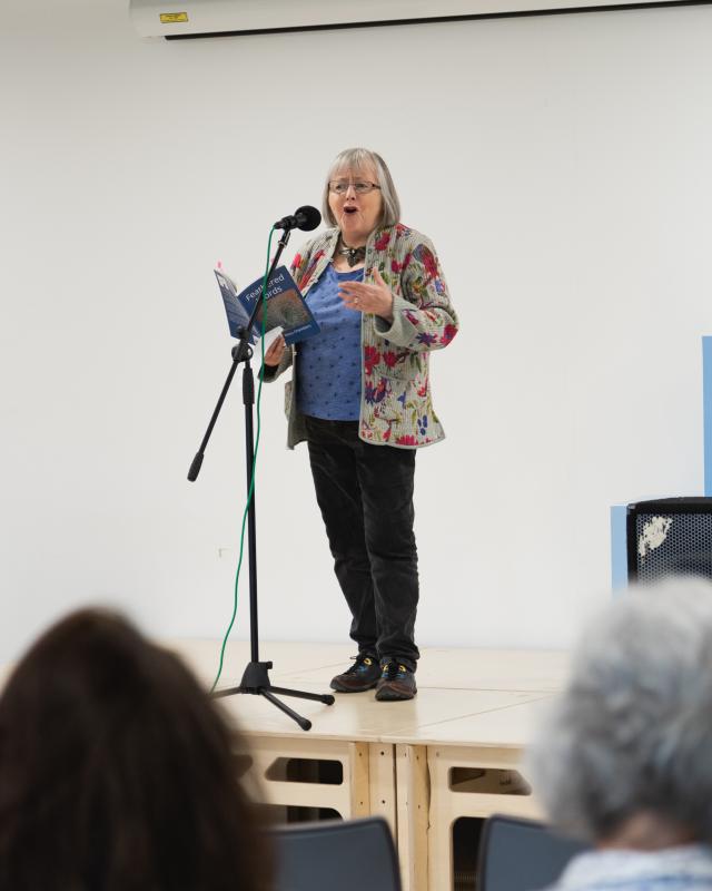 Woman on stage with floral jacket, reading poetry into microphone 
