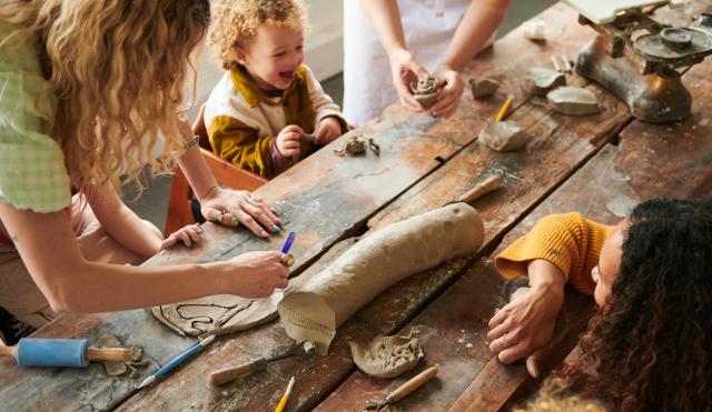 A wooden table, with clay items on the table. A child with two women adults playing with the clay, using clay tools. 