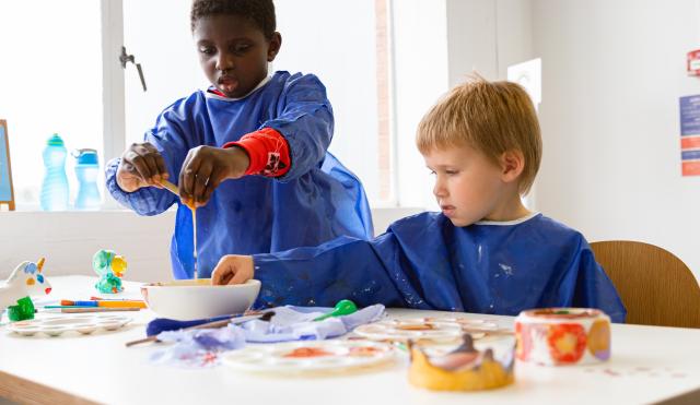 Two boys at a white table wearing blue aprons, playing with paint.