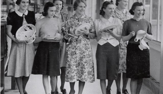 Black and white photograph of 7 female factory workers walking with plates in hand