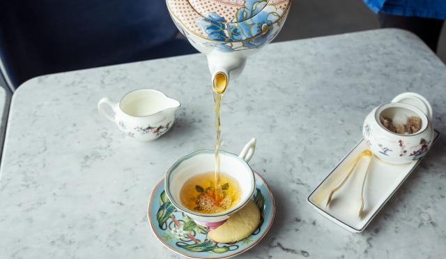 Image of floral Wedgwood Tea Pot pouring a loose leaf tea into a tea cup and saucer. Also to the right hand side of the table sits a dish with a sugar pot on and gold tongs. A matching milk jug is on the left hand side of the image.