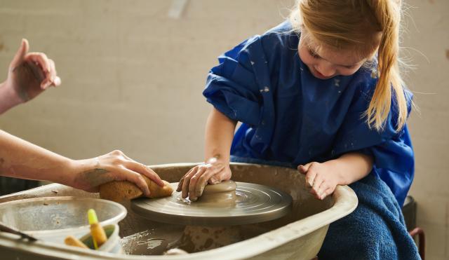 Girl is sitting at a potters wheel with a blue apron and squashing down the clay on the wheel top